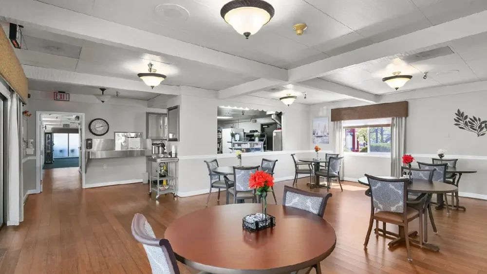 A dining area with wooden floors, several tables and chairs, and a pass-through opening to a kitchen area in the back
