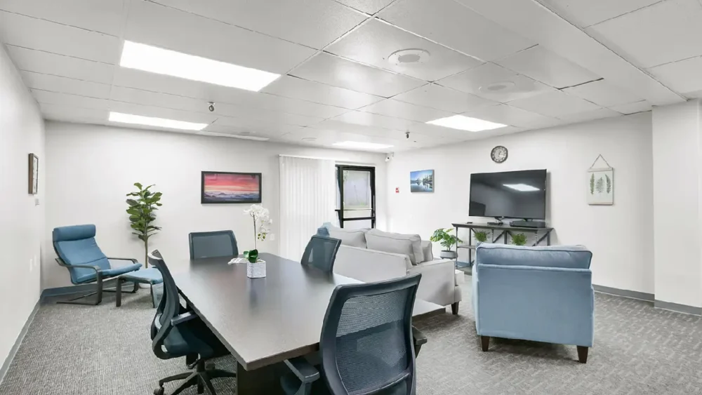 A meeting or conference room with a large dark table and rolling chairs, a light gray couch, and two light blue armchairs, with a television mounted on the wall