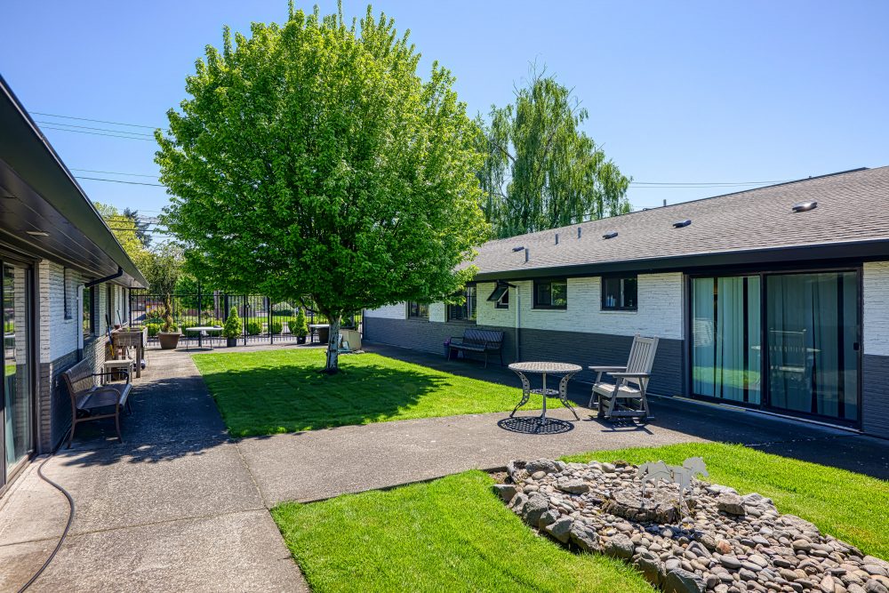 A grassy courtyard with a large tree, a small table with two chairs, a bench, and two buildings with black and white siding